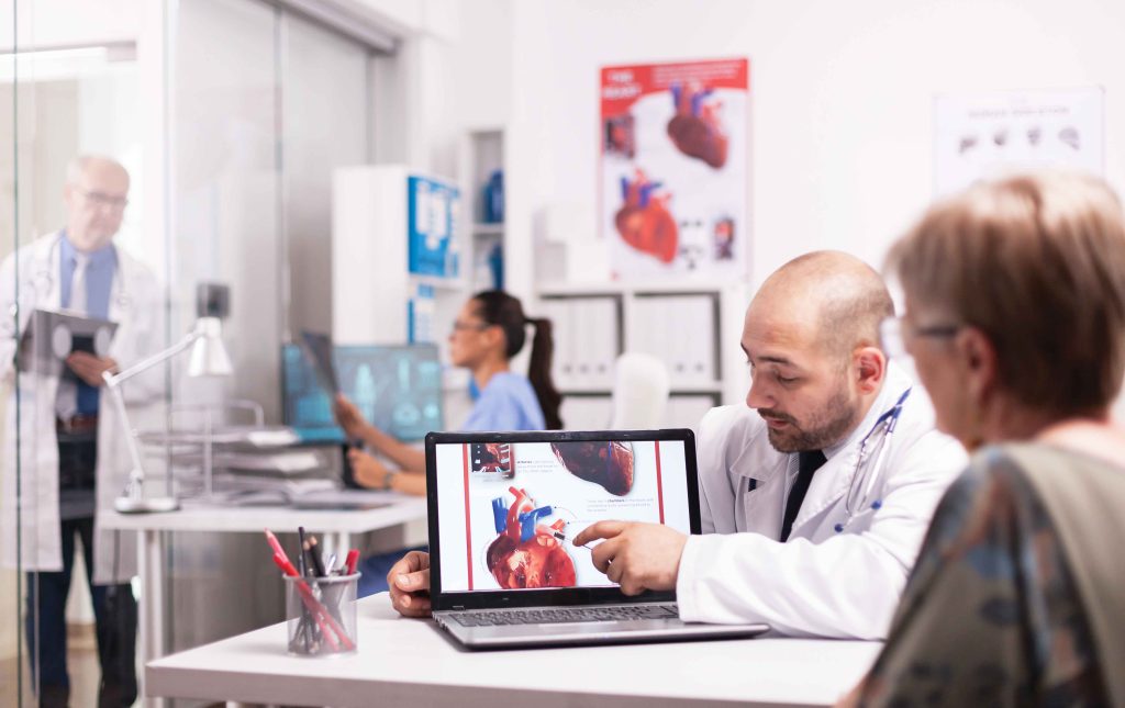 A stock image of a medical office with a man in a white medical coat pointing to an image of a diagram of the heart on the screen of a laptop. A woman in blue scrubs works at a computer in the background.