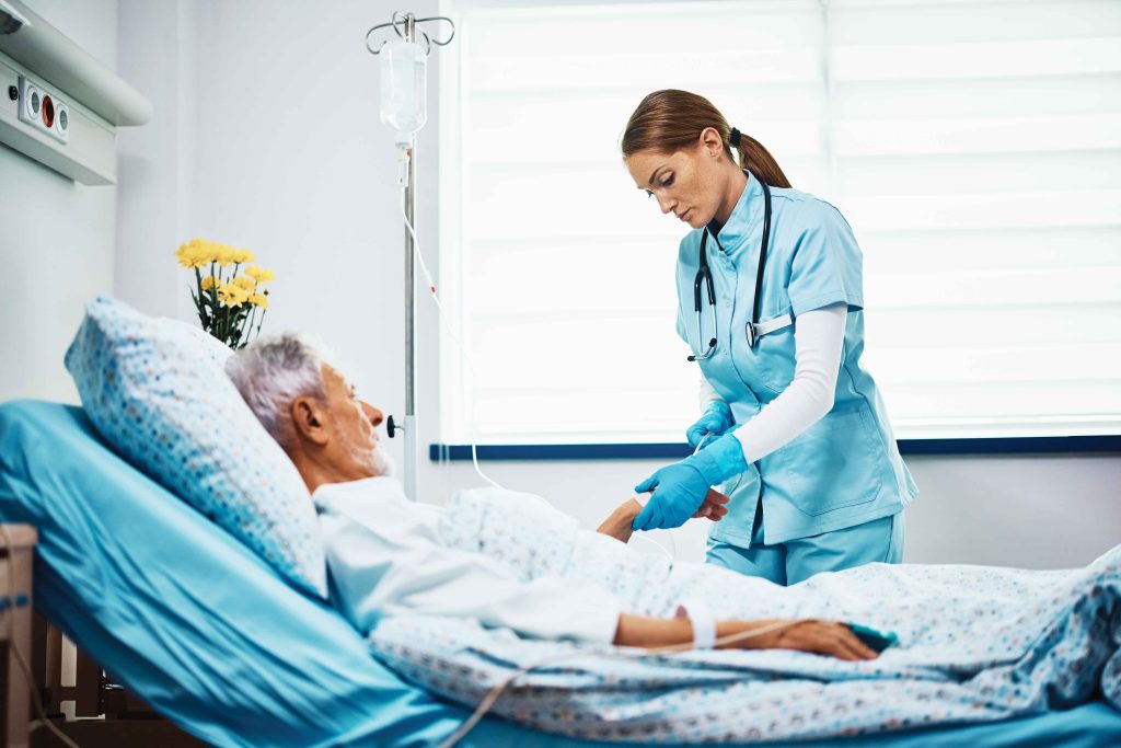 An older white man with gray hair lies in a hospital bed while a woman in blue scrubs lifts his left wrist to administer an IV.