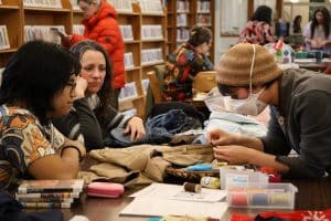 A woman wearing a face mask demonstrates how to stitch clothing to a young man and woman.