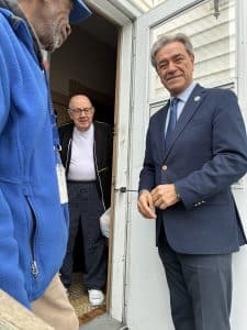An older man stands in the doorway of his home holding the meal that has just been delivered from a man in a suit who is looking at the camera.