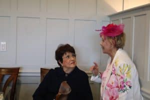 Seated older woman speaks to another older woman at an event table.