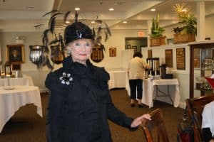 Older woman dress well in black with a fancy black hat with feathers smiles at camera.